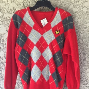 May include: Red V-neck sweater with a gray and black argyle pattern. The sweater has long sleeves and a small yellow bird emblem on the chest. A tag hangs from the neckline.