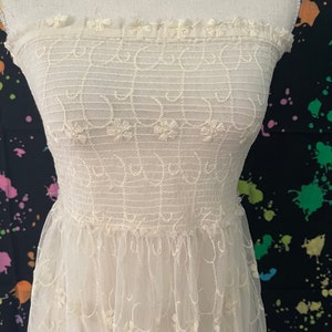 Pu&ograve; includere: Abito senza spalline bianco sporco con ricami floreali e gonna in tulle a strati. Il corpetto presenta linee orizzontali e dettagli floreali, mentre la gonna ha delicati dettagli ricamati. L'abito &egrave; esposto su un manichino.