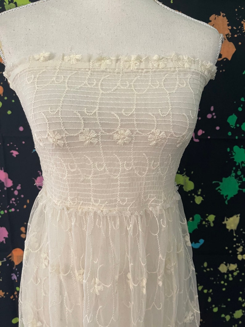 Pu&ograve; includere: Abito senza spalline bianco sporco con ricami floreali e gonna in tulle a strati. Il corpetto presenta linee orizzontali e dettagli floreali, mentre la gonna ha delicati dettagli ricamati. L'abito &egrave; esposto su un manichino.