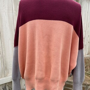 May include: A colour block sweater in shades of burgundy, peach, and grey. The sweater has a ribbed collar and cuffs, with a relaxed fit. The sleeves are grey, and the body is peach and burgundy.