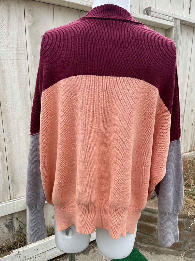 May include: A colour block sweater in shades of burgundy, peach, and grey. The sweater has a ribbed collar and cuffs, with a relaxed fit. The sleeves are grey, and the body is peach and burgundy.