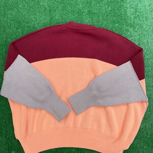 May include: A colour-block sweater in a relaxed fit. The top section is burgundy, the middle is peach, and the sleeves are grey. The sweater has a ribbed knit texture and a crew neck.