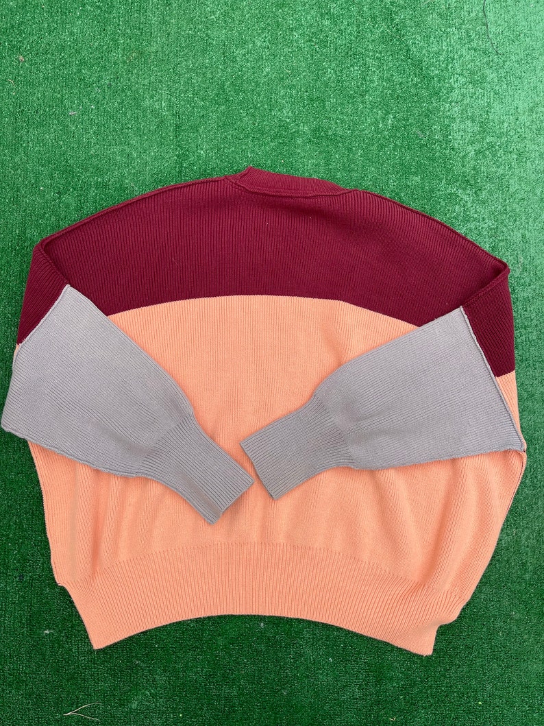 May include: A colour-block sweater in a relaxed fit. The top section is burgundy, the middle is peach, and the sleeves are grey. The sweater has a ribbed knit texture and a crew neck.