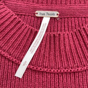 May include: Close-up of a burgundy ribbed knit sweater. The sweater has a crew neck and a small metal tag that reads "Free People". A white tag with text is attached to the sweater.