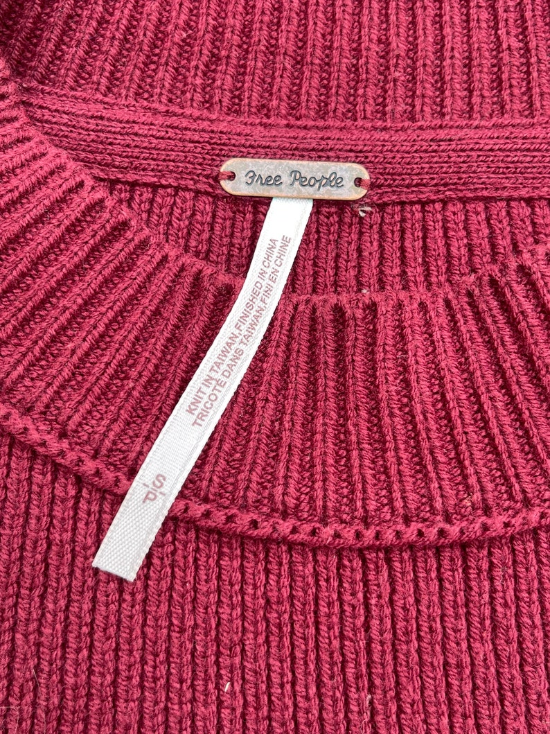 May include: Close-up of a burgundy ribbed knit sweater. The sweater has a crew neck and a small metal tag that reads "Free People". A white tag with text is attached to the sweater.