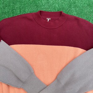 May include: A colour-block sweater in burgundy, peach, and grey. The ribbed knit sweater has a crew neck and long sleeves. The sleeves are grey, the body is peach, and the top is burgundy.