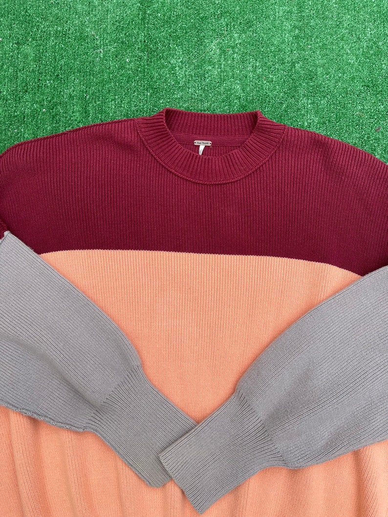 May include: A colour-block sweater in burgundy, peach, and grey. The ribbed knit sweater has a crew neck and long sleeves. The sleeves are grey, the body is peach, and the top is burgundy.