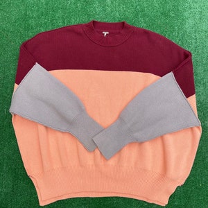 May include: A colour block sweater in burgundy, peach, and grey. The sweater has a ribbed texture and a crew neck. The sleeves are grey and flared at the wrist. The sweater is displayed on a green surface.