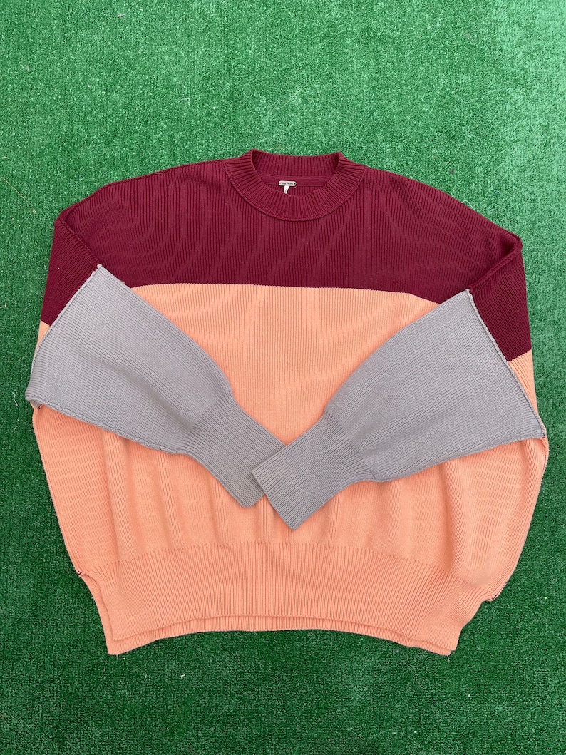 May include: A colour block sweater in burgundy, peach, and grey. The sweater has a ribbed texture and a crew neck. The sleeves are grey and flared at the wrist. The sweater is displayed on a green surface.