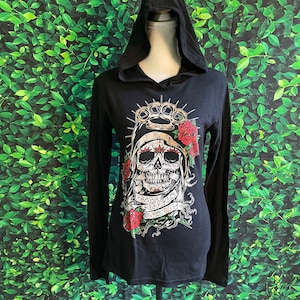 May include: Black hooded long-sleeve shirt featuring a skull graphic with brass knuckles, roses, and the text "Brass Knuckle Bitch". The shirt is displayed on a mannequin against a green leafy background.