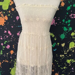 Pu&ograve; includere: Abito senza spalline bianco sporco con ricami floreali. L'abito presenta un corpetto aderente e una gonna lunga e fluida. Il tessuto sembra essere un delicato pizzo o tulle, con una trama sottile. L'abito &egrave; esposto su un manichino.
