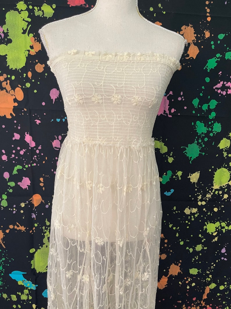Pu&ograve; includere: Abito senza spalline bianco sporco con ricami floreali. L'abito presenta un corpetto aderente e una gonna lunga e fluida. Il tessuto sembra essere un delicato pizzo o tulle, con una trama sottile. L'abito &egrave; esposto su un manichino.