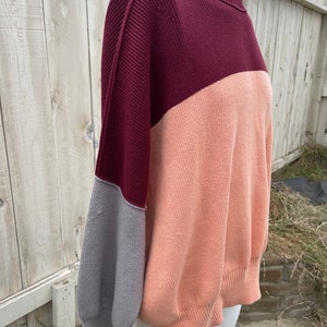 May include: A colour-blocked sweater in shades of burgundy, peach, and grey. The sweater has a ribbed neckline and cuffs, with a relaxed fit. The sleeves are a contrasting grey colour, adding a unique design element to the garment.