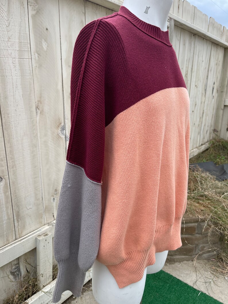 May include: A colour-blocked sweater in shades of burgundy, peach, and grey. The sweater has a ribbed neckline and cuffs, with a relaxed fit. The sleeves are a contrasting grey colour, adding a unique design element to the garment.