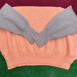 May include: A colour-block sweater in a ribbed knit design. The sweater features a burgundy top, a peach middle section, and grey sleeves. The sleeves have a ribbed cuff. The sweater is made of a soft, comfortable material.