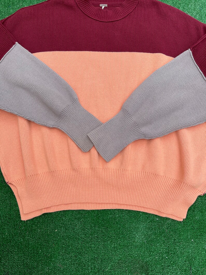 May include: A colour-block sweater in a ribbed knit design. The sweater features a burgundy top, a peach middle section, and grey sleeves. The sleeves have a ribbed cuff. The sweater is made of a soft, comfortable material.