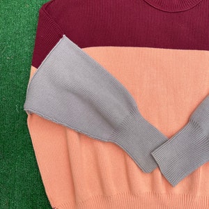 May include: A colour-block sweater in a ribbed knit design. The sweater features a burgundy top, a peach middle section, and grey sleeves. The sweater has a crew neck and a relaxed fit, suitable for casual wear.