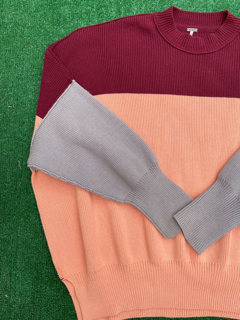 May include: A colour-block sweater in a ribbed knit design. The sweater features a burgundy top, a peach middle section, and grey sleeves. The sweater has a crew neck and a relaxed fit, suitable for casual wear.