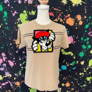 May include: Tan short-sleeve t-shirt with horizontal brown and gray stripes. The shirt features a colorful patch of a cartoon bulldog wearing a red hat. A vintage doll head is placed on top of the mannequin.