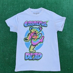 May include: White t-shirt featuring a tie-dye dancing bear graphic with the words "Grateful Dead" in a colorful, bubble-style font. The bear is set against a light blue circle. The shirt is made by Liquid Blue.