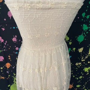 Pu&ograve; includere: Un abito color crema senza spalline con ricami floreali intricati. Il corpetto presenta un motivo dettagliato di riccioli ricamati e piccoli fiori, mentre la gonna &egrave; realizzata in un tessuto trasparente e fluido. L'abito &egrave; esposto su un manichino.