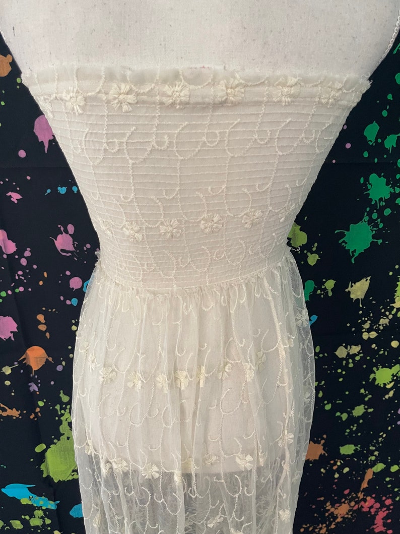 Pu&ograve; includere: Un abito color crema senza spalline con ricami floreali intricati. Il corpetto presenta un motivo dettagliato di riccioli ricamati e piccoli fiori, mentre la gonna &egrave; realizzata in un tessuto trasparente e fluido. L'abito &egrave; esposto su un manichino.