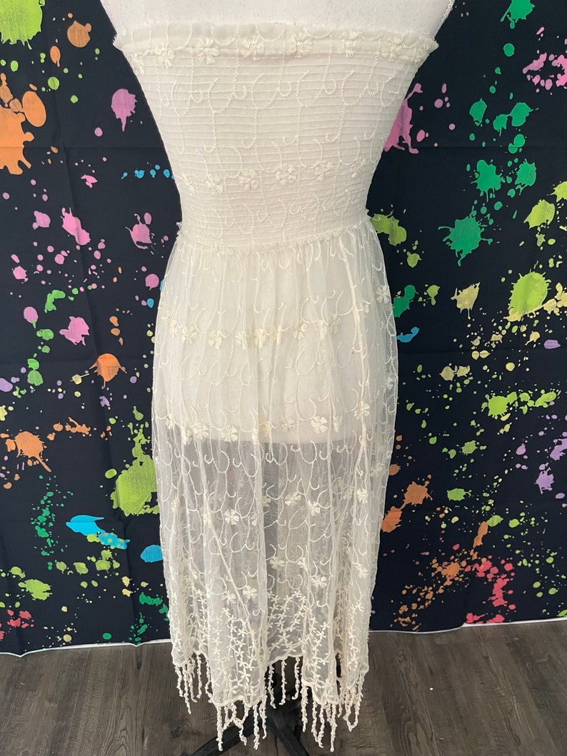 Pu&ograve; includere: Un abito senza spalline color crema con ricami floreali intricati. L'abito presenta un corpetto aderente e una gonna fluida e trasparente che arriva a met&agrave; polpaccio, rifinita con un orlo con frange. Il tessuto &egrave; delicato e leggero.