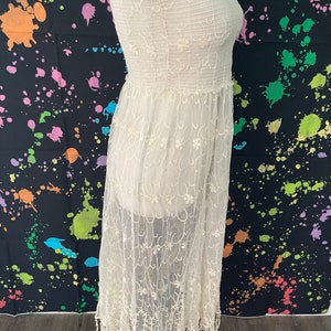 Pu&ograve; includere: Abito senza spalline color crema con sovrapposizione di pizzo floreale. L'abito presenta un corpetto a punto smock e una gonna fluida con orlo a frange. L'abito &egrave; esposto su un manichino su sfondo nero con schizzi di vernice colorati.
