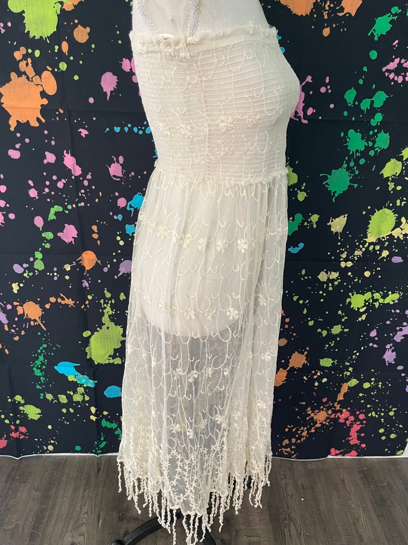 Pu&ograve; includere: Abito senza spalline color crema con sovrapposizione di pizzo floreale. L'abito presenta un corpetto a punto smock e una gonna fluida con orlo a frange. L'abito &egrave; esposto su un manichino su sfondo nero con schizzi di vernice colorati.
