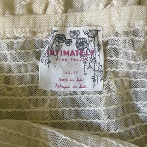 Pu&ograve; includere: Primo piano di un capo in pizzo color crema. Il tessuto ha un aspetto delicato e strutturato con motivi intricati. Un'etichetta con le parole "Intimately Free People" &egrave; visibile, insieme a "Made in India" e "Fabriqu&eacute; en Inde".