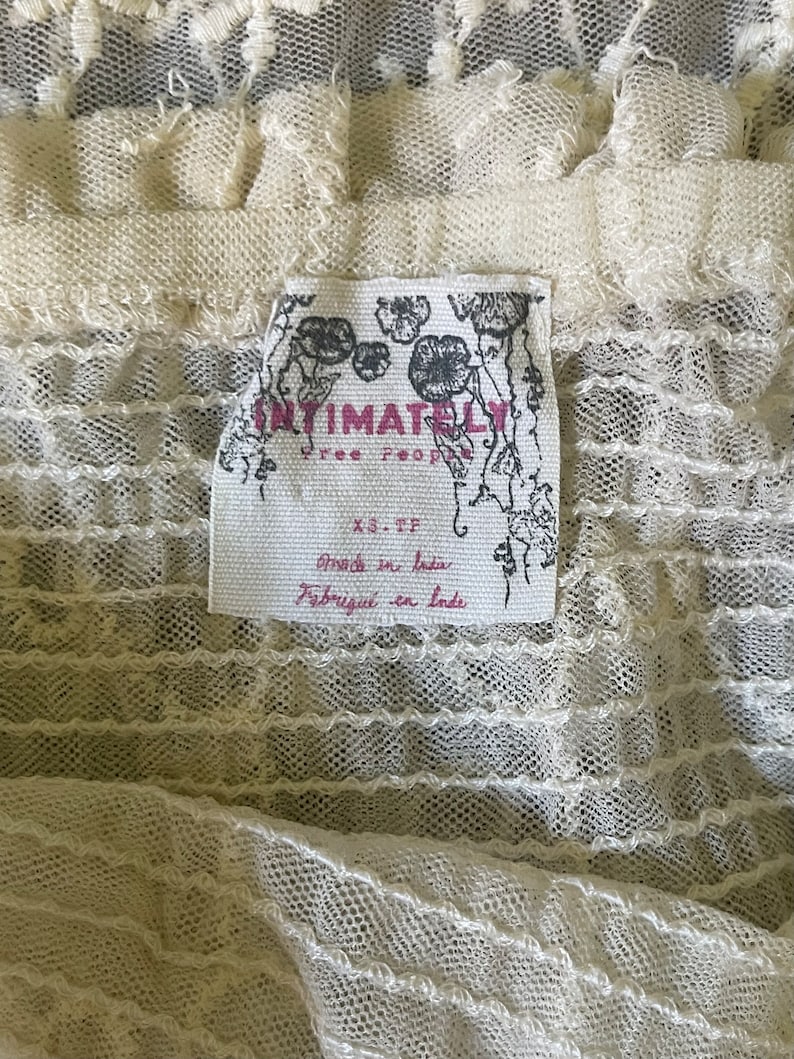 Pu&ograve; includere: Primo piano di un capo in pizzo color crema. Il tessuto ha un aspetto delicato e strutturato con motivi intricati. Un'etichetta con le parole "Intimately Free People" &egrave; visibile, insieme a "Made in India" e "Fabriqu&eacute; en Inde".