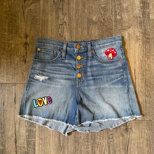 May include: Blue denim shorts with a high-rise waist, a button-up fly, and a frayed hem. The shorts feature a red and white mushroom patch on the right pocket and a colorful patch on the left leg that displays the word LOVE in block letters.
