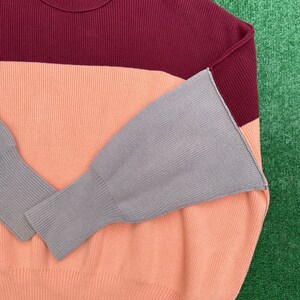 May include: A colour-block sweater in shades of burgundy, peach, and grey. The sweater features a ribbed texture and a crew neck. The sleeves are a contrasting grey colour. The brand name "Free People" is visible on the neckline.