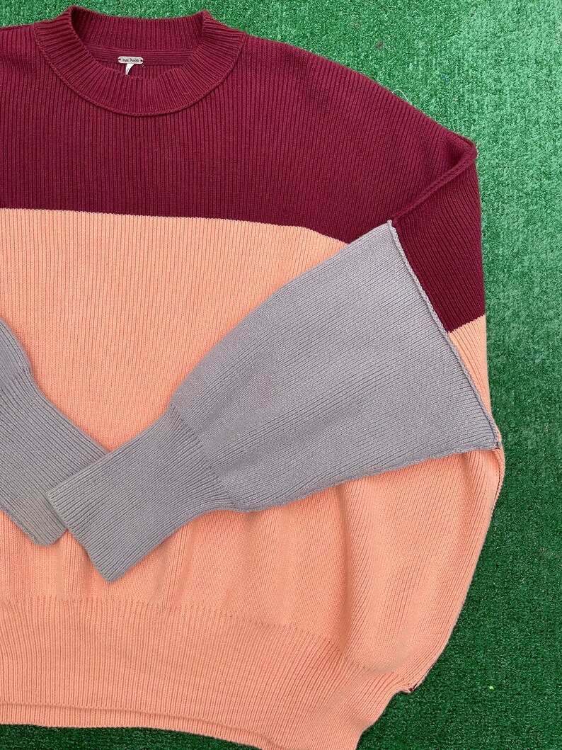 May include: A colour-block sweater in shades of burgundy, peach, and grey. The sweater features a ribbed texture and a crew neck. The sleeves are a contrasting grey colour. The brand name "Free People" is visible on the neckline.