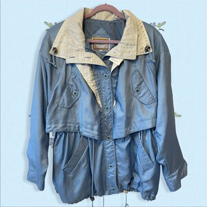 May include: A light blue jacket with a beige collar and accents. The jacket has a drawstring waist, button closures, and pockets. The jacket is made of a shiny material and has a vintage style.