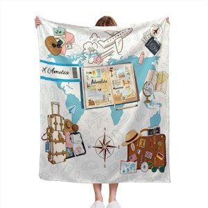 May include: A white fleece blanket featuring a travel-themed print with a light blue world map background. Illustrations include a vintage suitcase, a compass, an open scrapbook labeled Adventure Memories, a plane, and a ticket with the name Amelia.