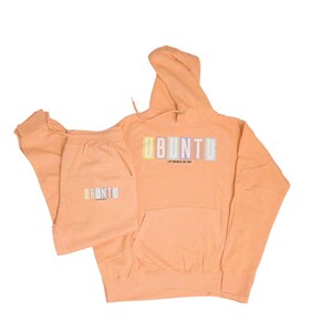 May include: Peach-colored Ubuntu hoodie and sweatpants set. The hoodie has a front pocket and a drawstring hood. The word "UBUNTU" is printed across the chest in pastel colors. The sweatpants have the word "UBUNTU" printed on the left leg.