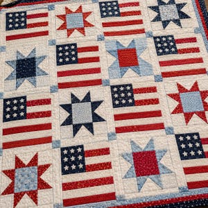 May include: A quilted textile featuring a repeating pattern of American flags and star shapes. The flags have red and white stripes with a blue field of white stars. The stars are in shades of blue and red, set against a white background.