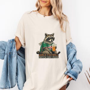 Może przedstawiać: Kremowa koszulka z szopem w okularach przeciwsłonecznych, szaliku i koszulce piłkarskiej, trzymającym napój. Koszulka ma napis "WORLD FOOTBALL MATCH DAY MODE" w czcionce.