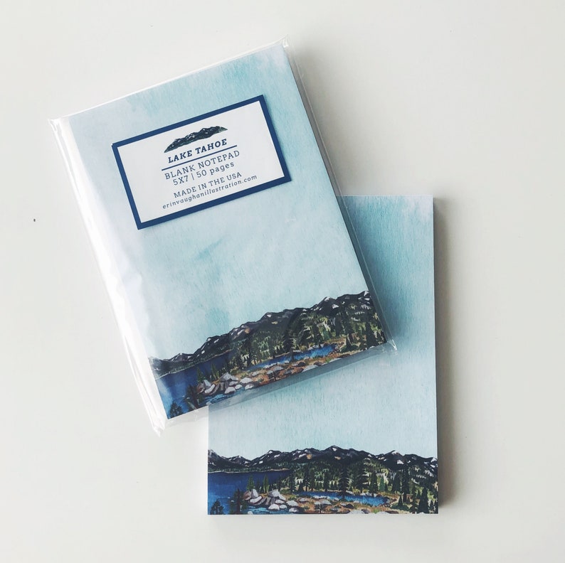 May include: Two blank notepads with a watercolor design. The design features a blue sky, a mountain range, and a lake. The notepads are labeled "Lake Tahoe Blank Notepad 5x7 | 50 pages". One notepad is in a clear plastic sleeve.
