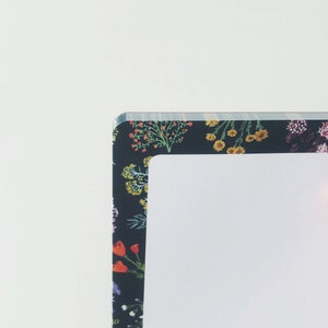 May include: A notebook with a floral pattern border. The border features a dark background with colorful flowers in shades of red, yellow, and purple. The notebook has a white interior.