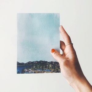 May include: A watercolor landscape print featuring a lake, mountains, and trees under a light blue sky. The artwork is held by a hand with orange nail polish and a silver ring. The print has a serene and natural theme.