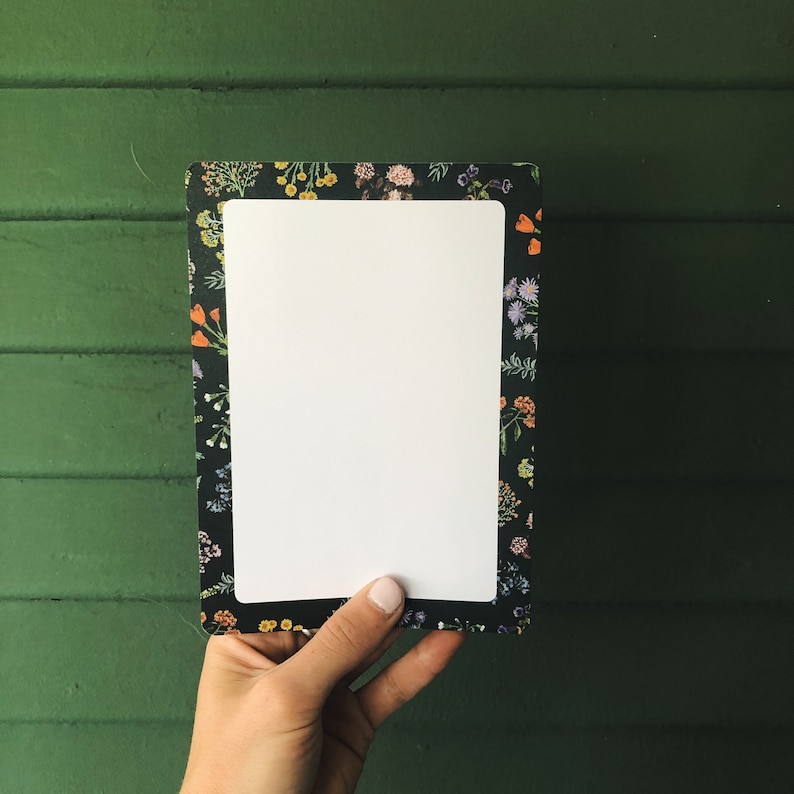 May include: Rectangular notepad with a white interior and a floral patterned border. The border features a dark background with colorful flowers in shades of orange, yellow, purple, and white. The notepad is held against a green background.