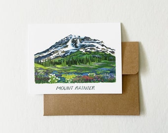 May include: A watercolor greeting card featuring Mount Rainier. The artwork depicts the mountain with snow-capped peaks, green forests, and a colorful wildflower meadow. The words "Mount Rainier" are printed at the bottom. The card is paired with a brown envelope.