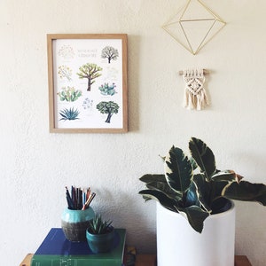 May include: A framed print titled "Native Plants & Joshua Tree" hangs on a white wall. Below, a wooden table holds a white ceramic pot with a plant, books, and a small teal vase with pencils. A gold geometric wall hanging and a macrame piece are also on the wall.