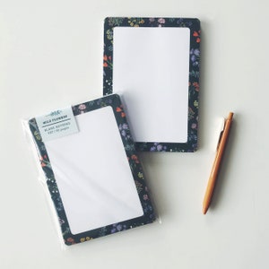 May include: Two blank notepads with a floral border and a brown pen on a white surface. One notepad is in a clear plastic sleeve. The notepads are 5x7 inches and have 50 pages. The floral border has a dark background with colorful flowers.