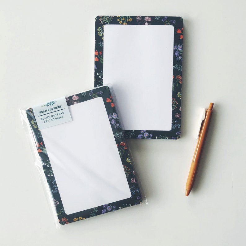 May include: Two blank notepads with a floral border and a brown pen on a white surface. One notepad is in a clear plastic sleeve. The notepads are 5x7 inches and have 50 pages. The floral border has a dark background with colorful flowers.