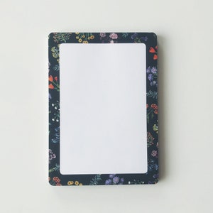 May include: Rectangular notepad with a white interior and a dark blue floral border. The border features a variety of colorful flowers, including red, yellow, and purple blooms. The notepad has rounded corners and a clean, modern design.