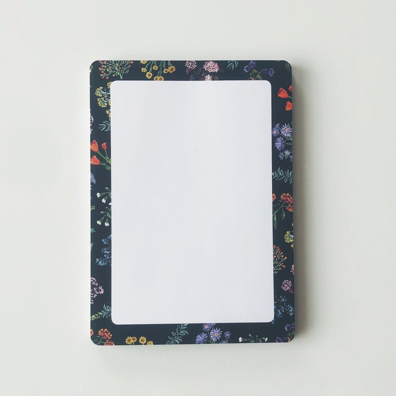 May include: Rectangular notepad with a white interior and a dark blue floral border. The border features a variety of colorful flowers, including red, yellow, and purple blooms. The notepad has rounded corners and a clean, modern design.