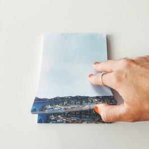 May include: A stack of landscape-themed notepads. The top notepad features a watercolor painting of a lake, mountains, and trees under a light blue sky. The notepad is held by a hand with orange nail polish.